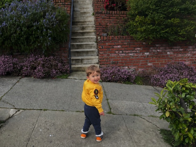 A toddler in a yellow jacket smiling back over his shoulder on a neighborhood sidewalk