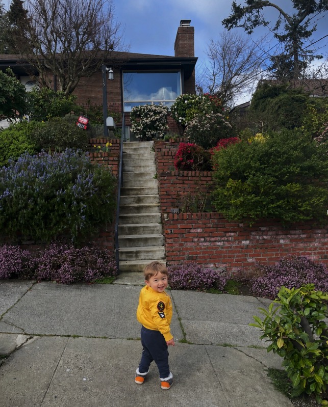 A toddler in a yellow jacket smiling back over his shoulder on a neighborhood sidewalk
